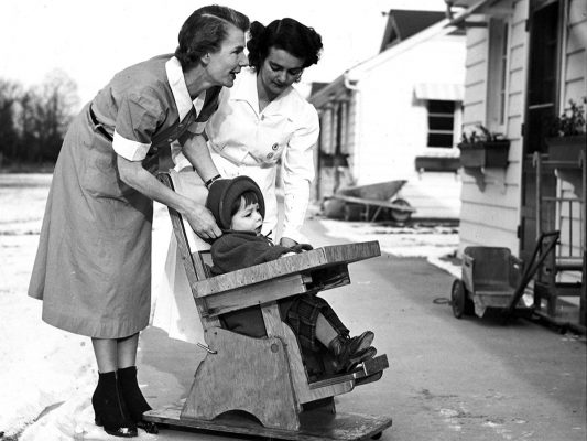 A black and white photo of two nurses pushing a girl in a wooden wheel chair