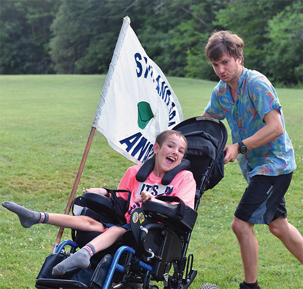 A councilor pushes a camper in her chair holding a camp flag