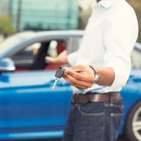 A man stands in front of a blue car holding out the keys.