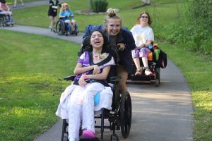 A councilor pushes a smiling girl in a wheel chair down a path at camp