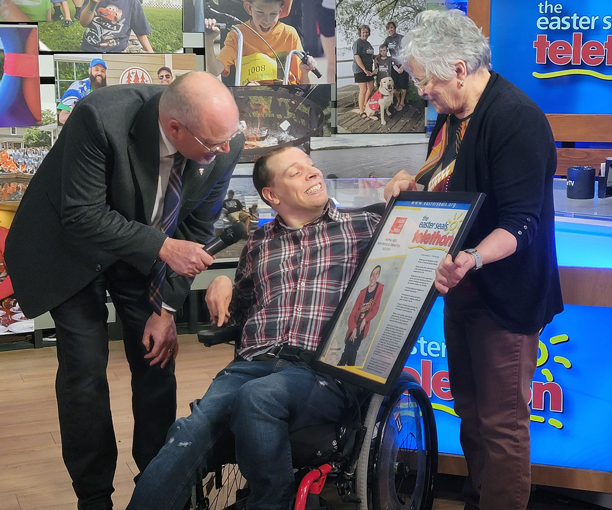 A young man in a wheelchair is presented a framed poster commemorating the Telethon.